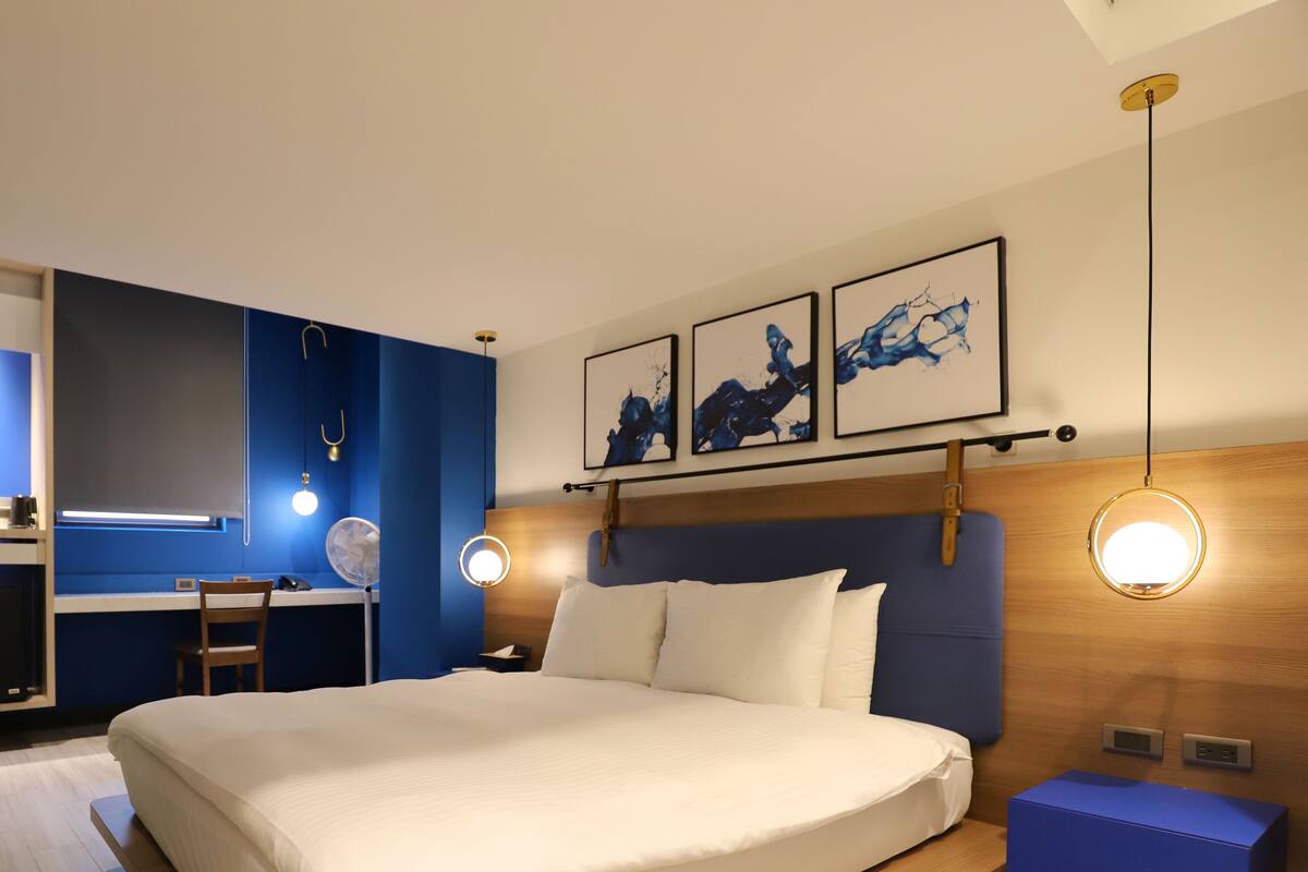 Habitación de hotel moderna con una cama grande, un escritorio y una silla. Cuenta con dos lámparas fijadas a la pared y un ventilador de techo.