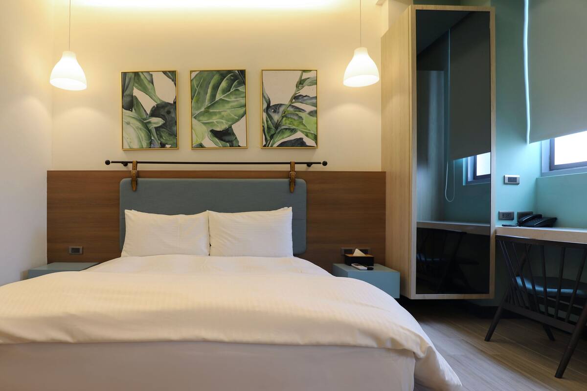 Una cama bien hecha con sábanas blancas, un cabecero de madera y dos mesitas de noche con lámparas. Encima de la cama, hay tres impresiones enmarcadas con motivos botánicos.