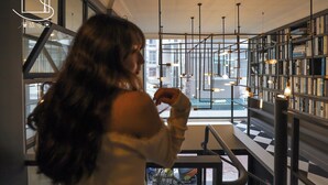 Une personne se tient près d’une fenêtre donnant sur une vue urbaine. L’intérieur, de style moderne, est caractérisé par un sol à carreaux noirs et blancs, une bibliothèque remplie de livres et des suspensions lumineuses qui pendent du plafond.