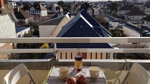 Un balcón con una mesa lista para dos personas, una botella y dos croissants. El balcón ofrece vista a una zona residencial con casas y una calle.