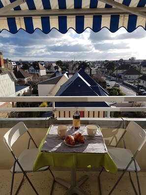 A balcony with a table set for two, a bottle, and two croissants. The balcony overlooks a residential area with houses and a street. - 3 * apartment at 200m BEACH & THALASSO - WIFI - BICYCLES (Ouistreham)