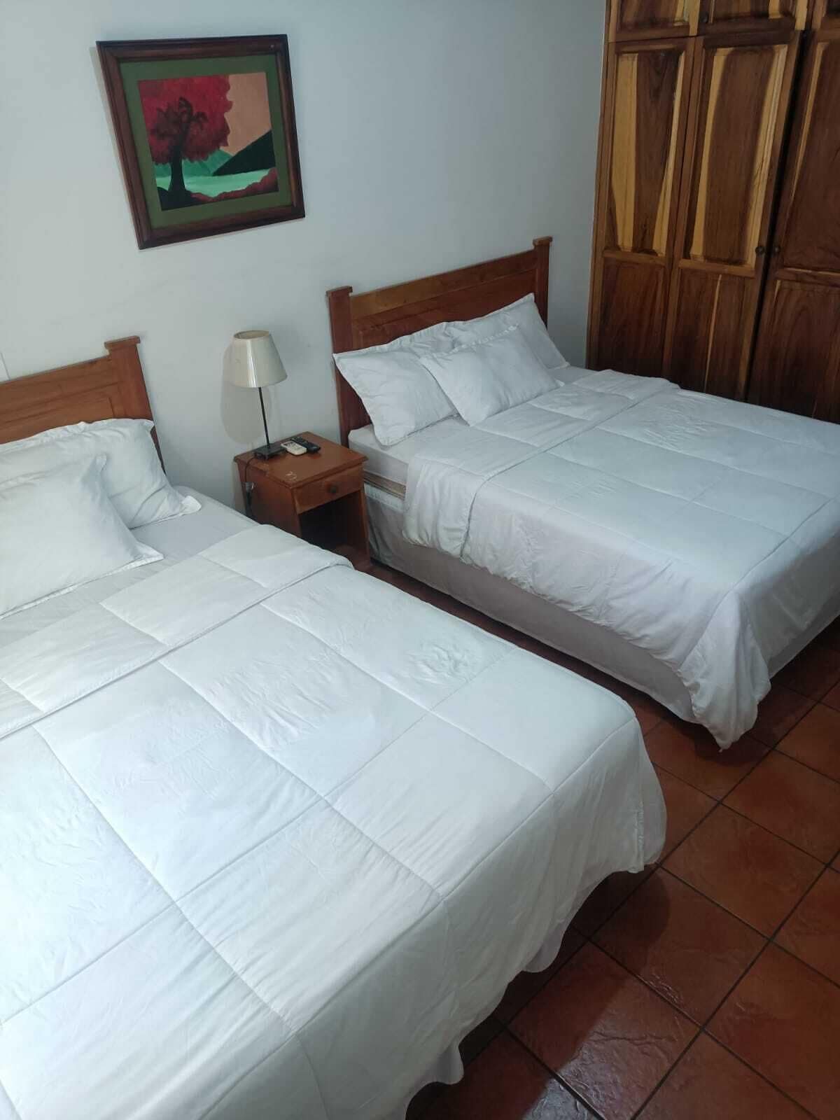 A hotel room with two single beds, a nightstand with a lamp, a wooden wardrobe, and a framed picture on the wall.