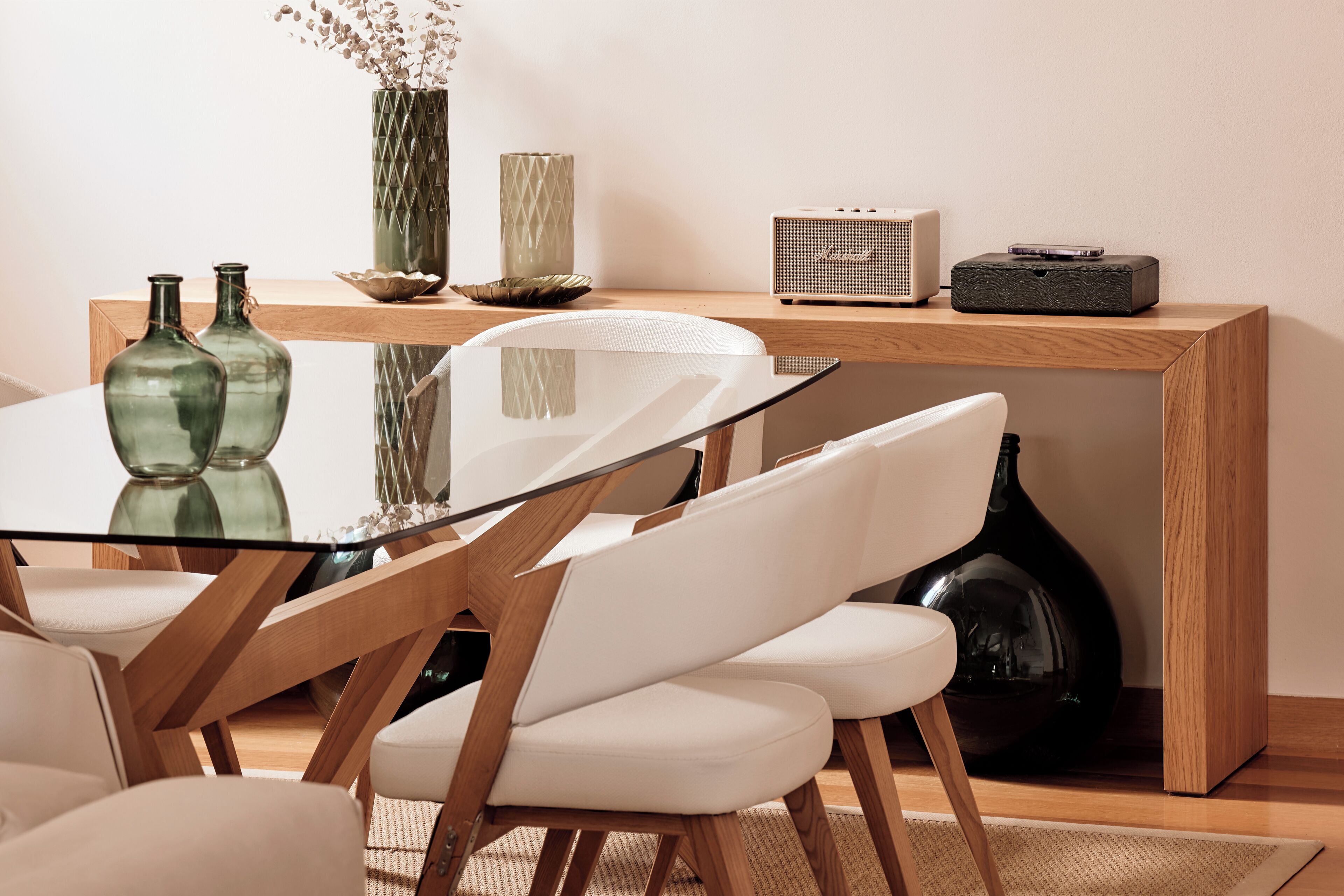 A modern dining area with a glass table, white chairs, and a wooden sideboard with decorative items and a speaker.