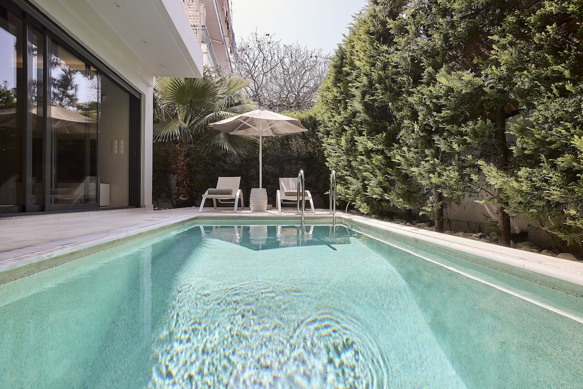 A swimming pool with clear water, surrounded by a modern building, a palm tree, and a well-maintained hedge.