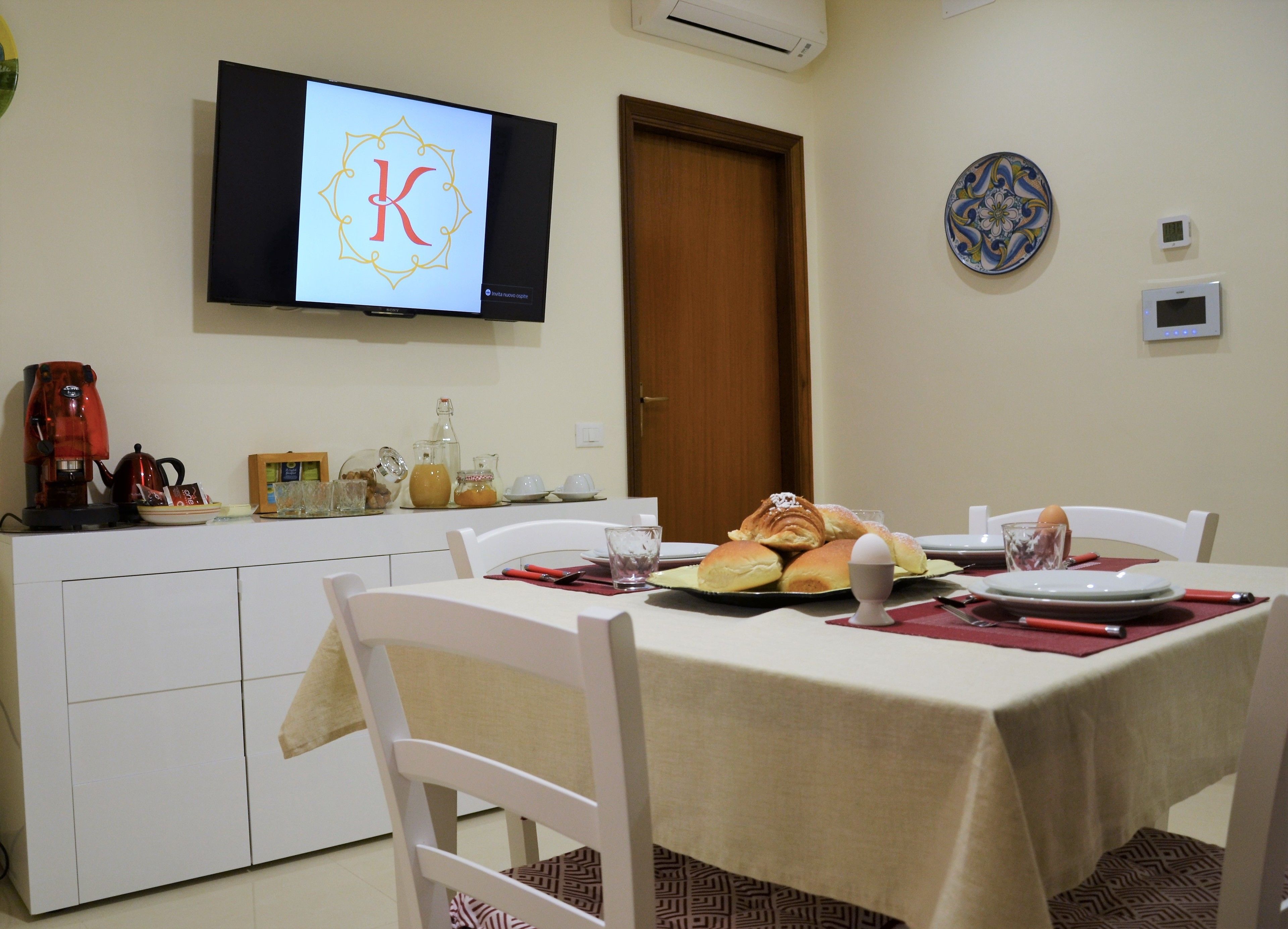 Un coin repas avec une table dressée pour deux personnes, un téléviseur affichant un logo et un espace cuisine équipé d’une machine à café et de pots.