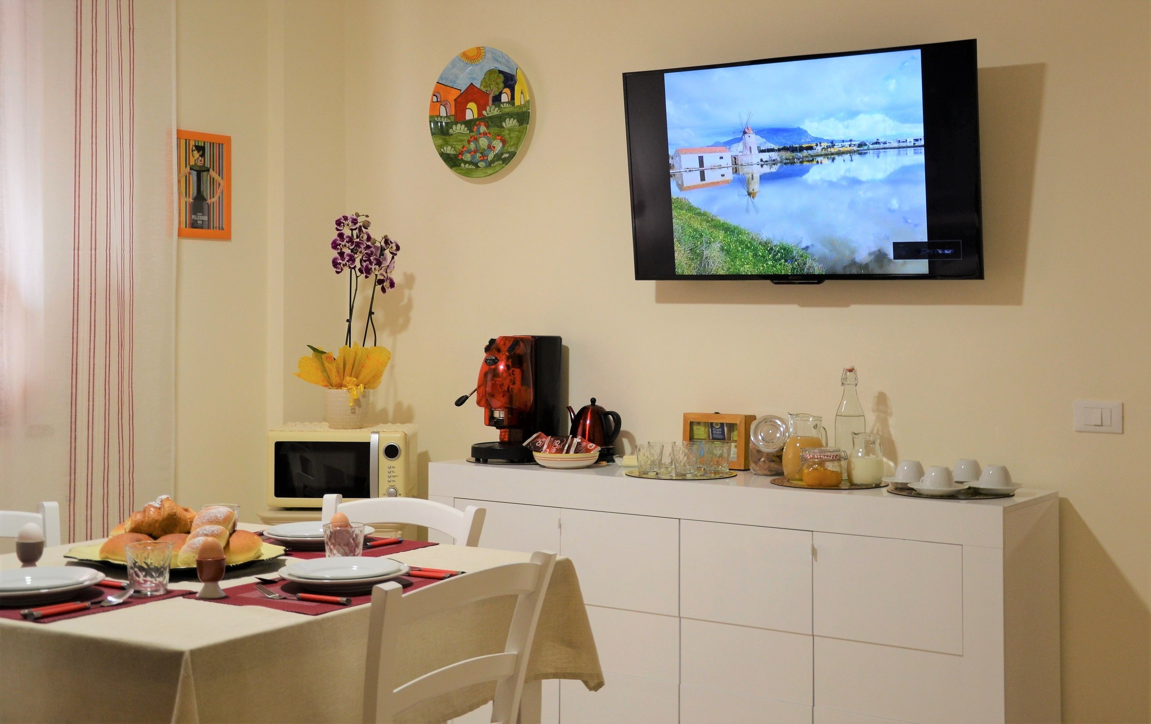Ein Essbereich mit an der Wand befestigtem Fernseher, der eine Landschaft zeigt, einer Kaffeemaschine, einem Wasserkocher und einem Tisch, gedeckt mit Tellern, Besteck und einer Blumenvase.