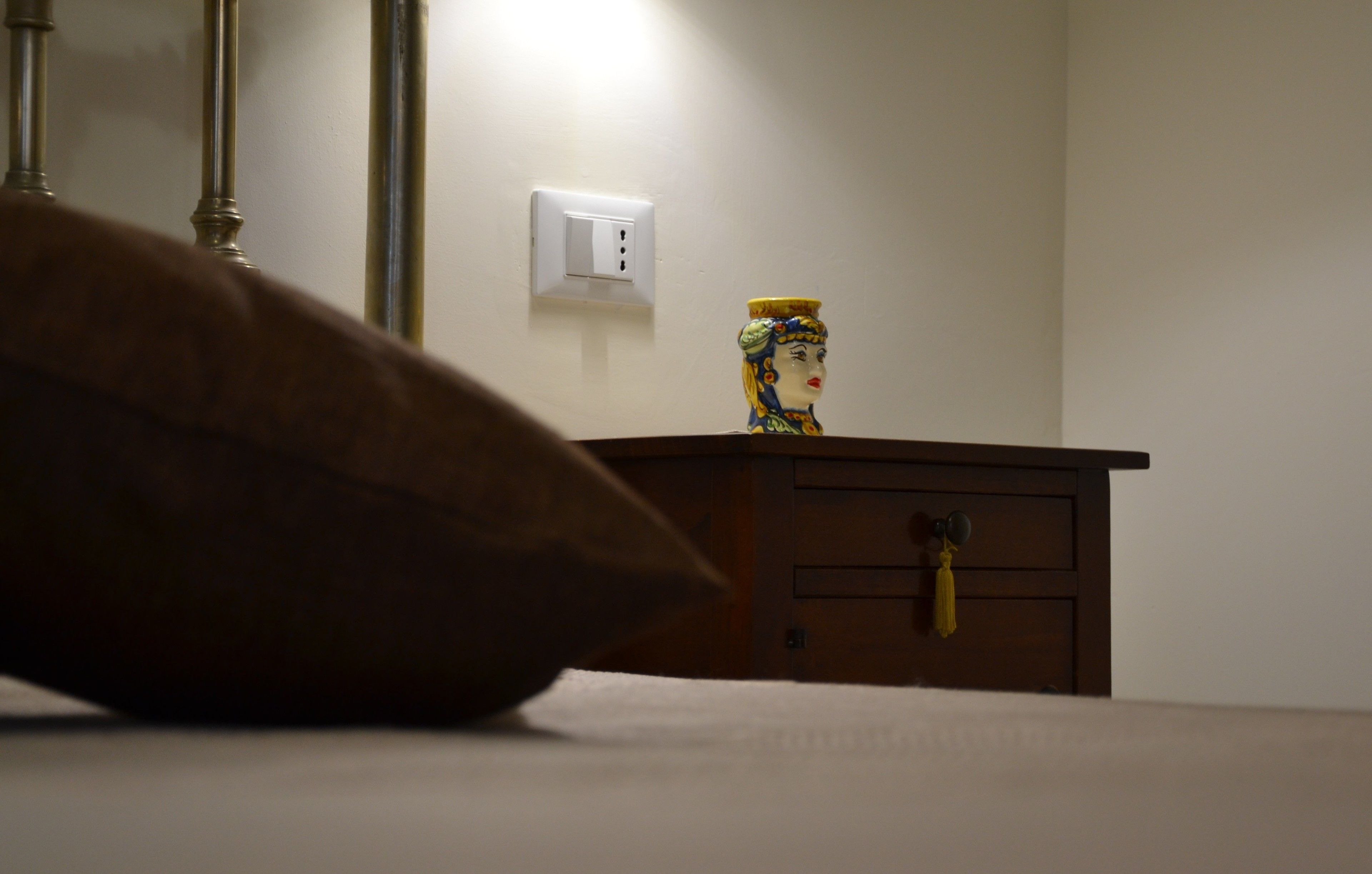 Un dormitorio con cabecera de madera, una mesita de noche con un florero decorativo y un enchufe de pared.