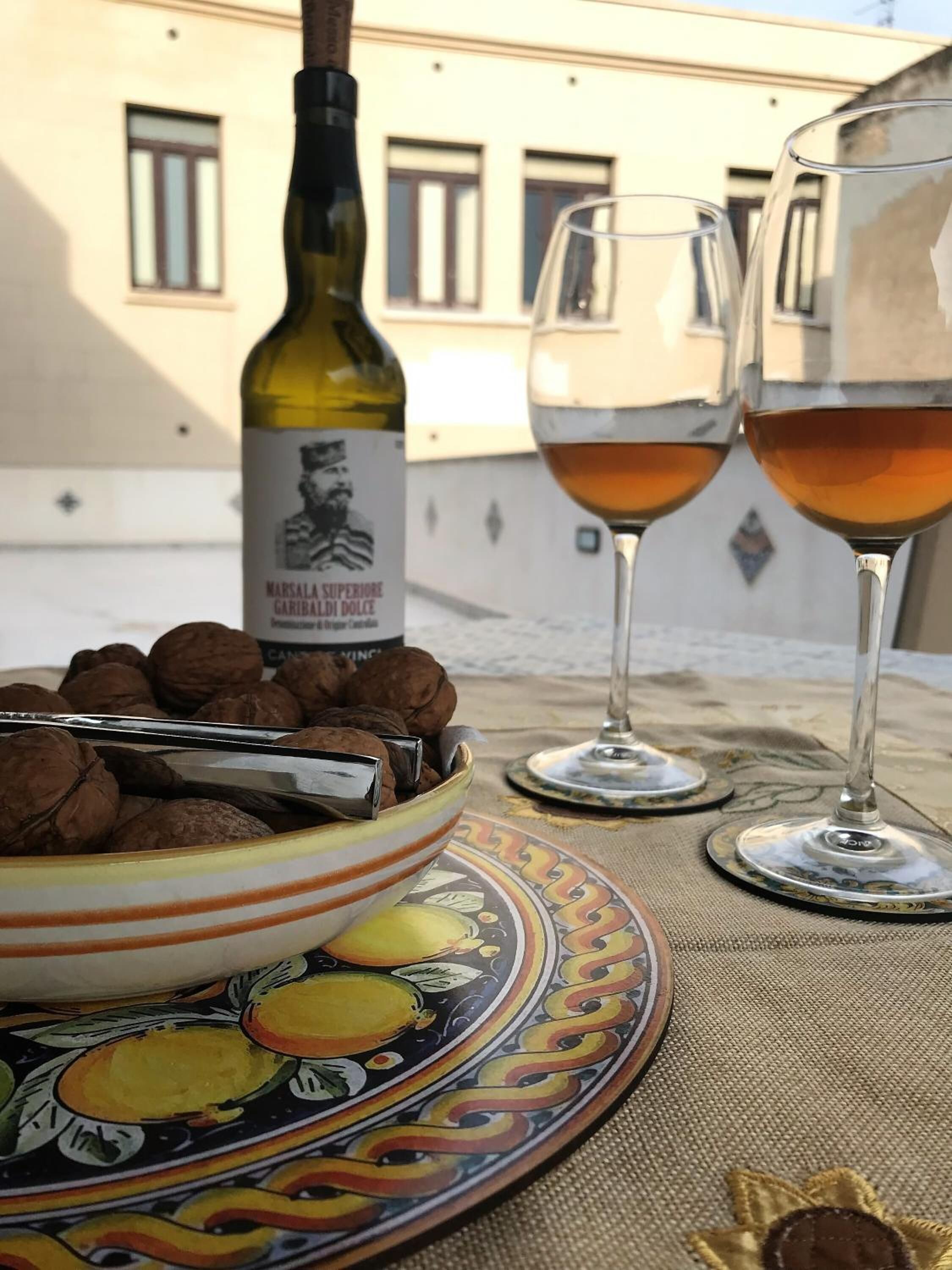 Un tavolo con una bottiglia di vino Marsala, due bicchieri da vino, una ciotola di noci e un piatto decorativo.