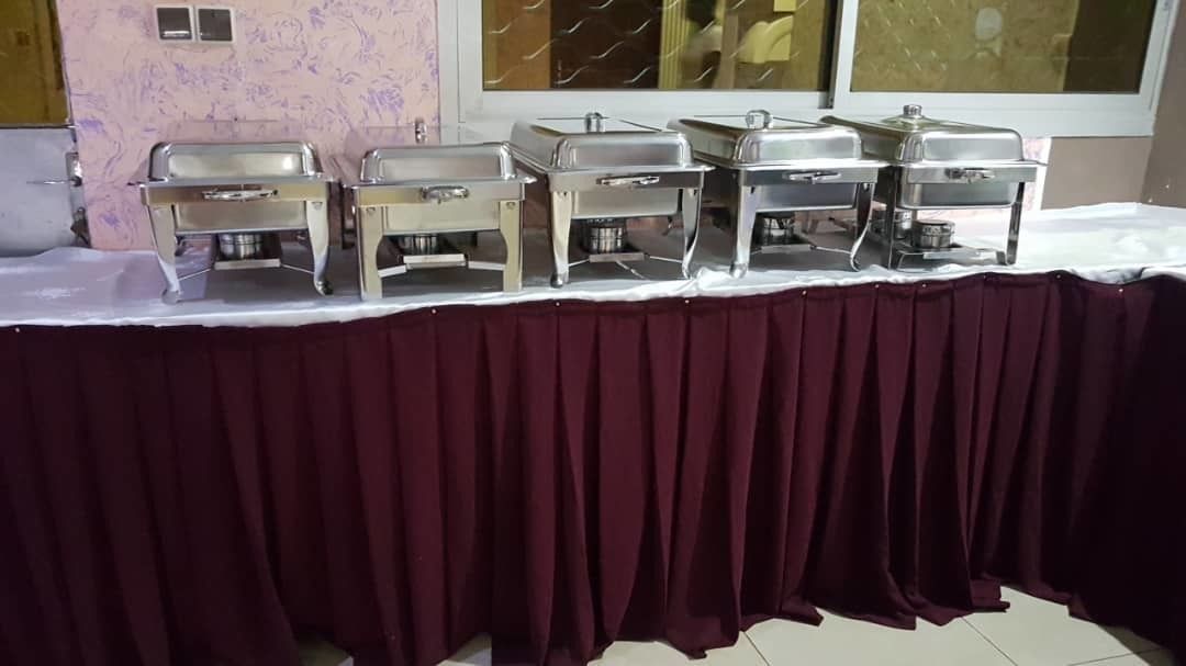 Un buffet composé de plusieurs plats de service en acier inoxydable, disposés sur une table recouverte d’une nappe blanche et agrémentée d’un chemin de table bordeaux.