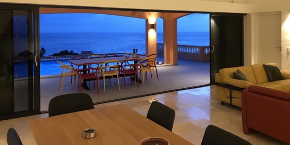 Une pièce dotée d’une grande porte-fenêtre coulissante donnant sur un balcon avec vue sur la mer. À l’intérieur, on trouve une table en bois avec des chaises et un canapé jaune.