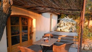 Una terraza de madera con una mesa y dos sillas de mimbre, una puerta de cristal y una estructura de techo de paja.