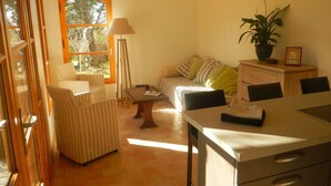 Una sala acogedora con un sofá, sillones a rayas, una mesa de centro y un escritorio con cajones. Se ve una ventana con vistas a árboles y una lámpara.