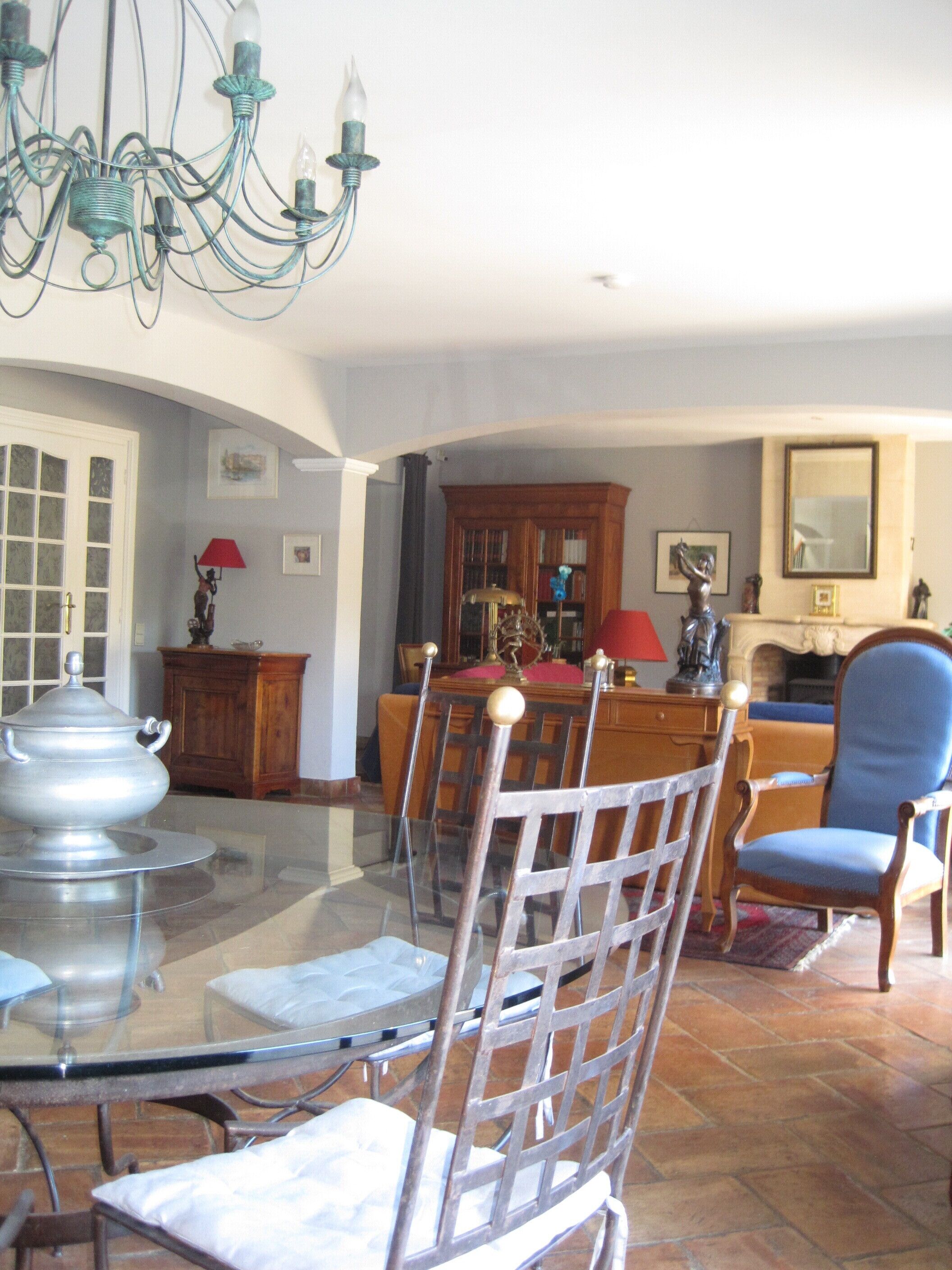 A dining area with a glass table, chairs, and a chandelier. There is a wooden cabinet and a red lamp in the background.