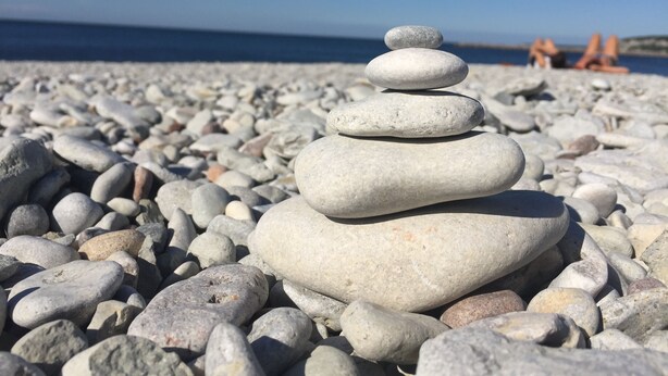 Een stapel gladde, ronde stenen op een kiezelstrand, met een helderblauwe lucht en een rustige zee op de achtergrond.