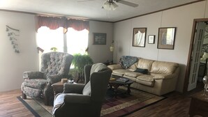 Un salon comprenant un canapé, un fauteuil et une table basse. Il y a une fenêtre avec des rideaux, un ventilateur de plafond et une porte qui donne sur une autre pièce.