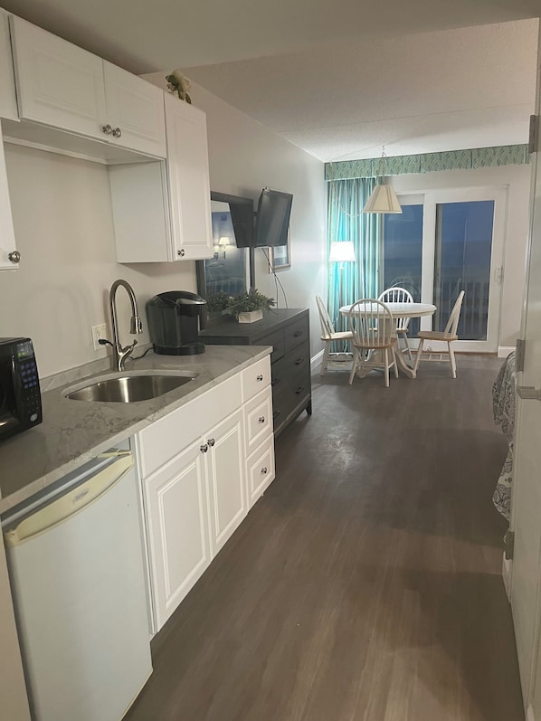 Cocina compacta con armarios blancos, microondas integrado y lavamanos. Hay una zona de comedor con mesa y sillas, y vistas al exterior a través de una puerta corrediza de vidrio.