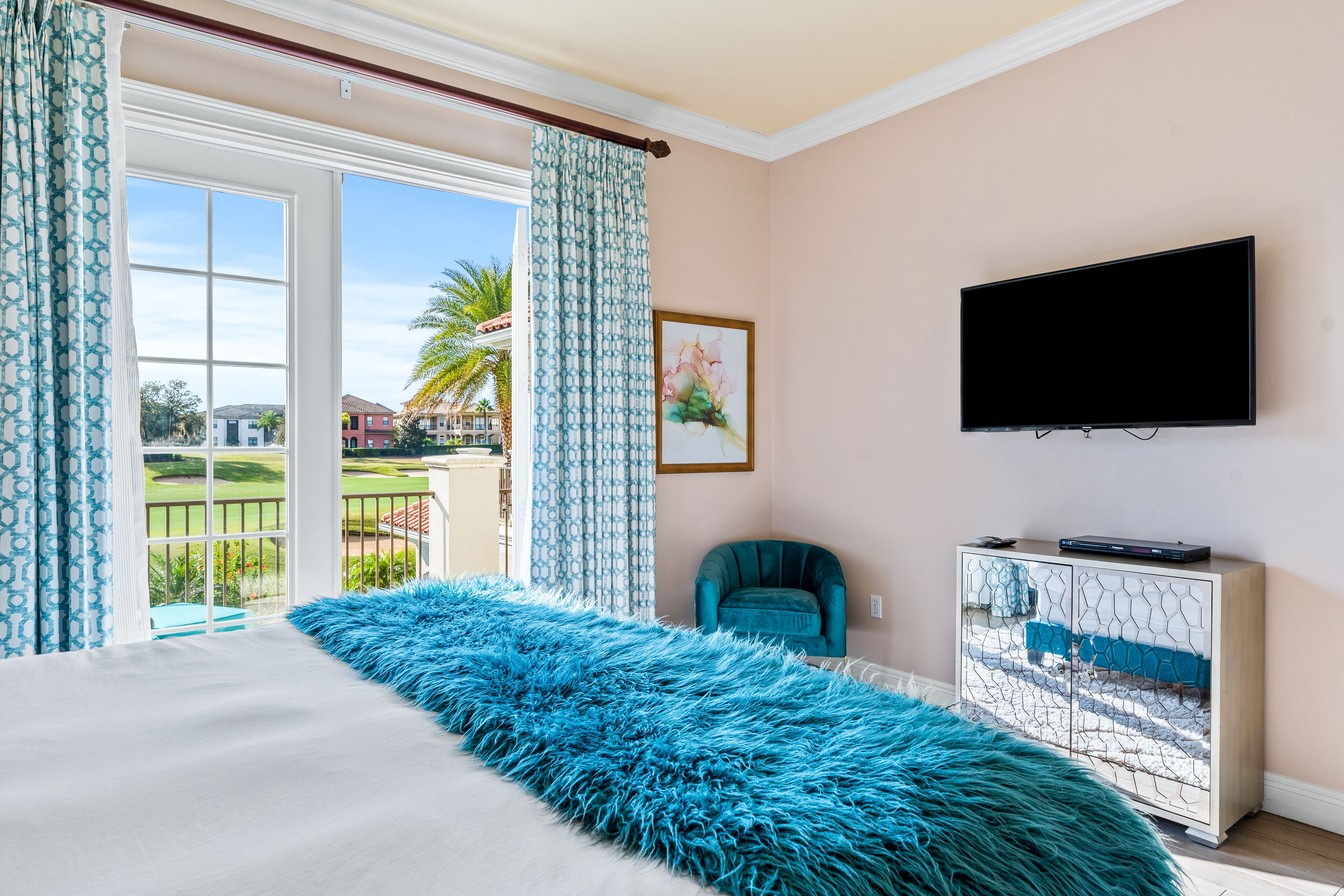 Une chambre avec un grand lit, un téléviseur fixé au mur, un fauteuil turquoise et une vue sur l’extérieur grâce à une porte coulissante.