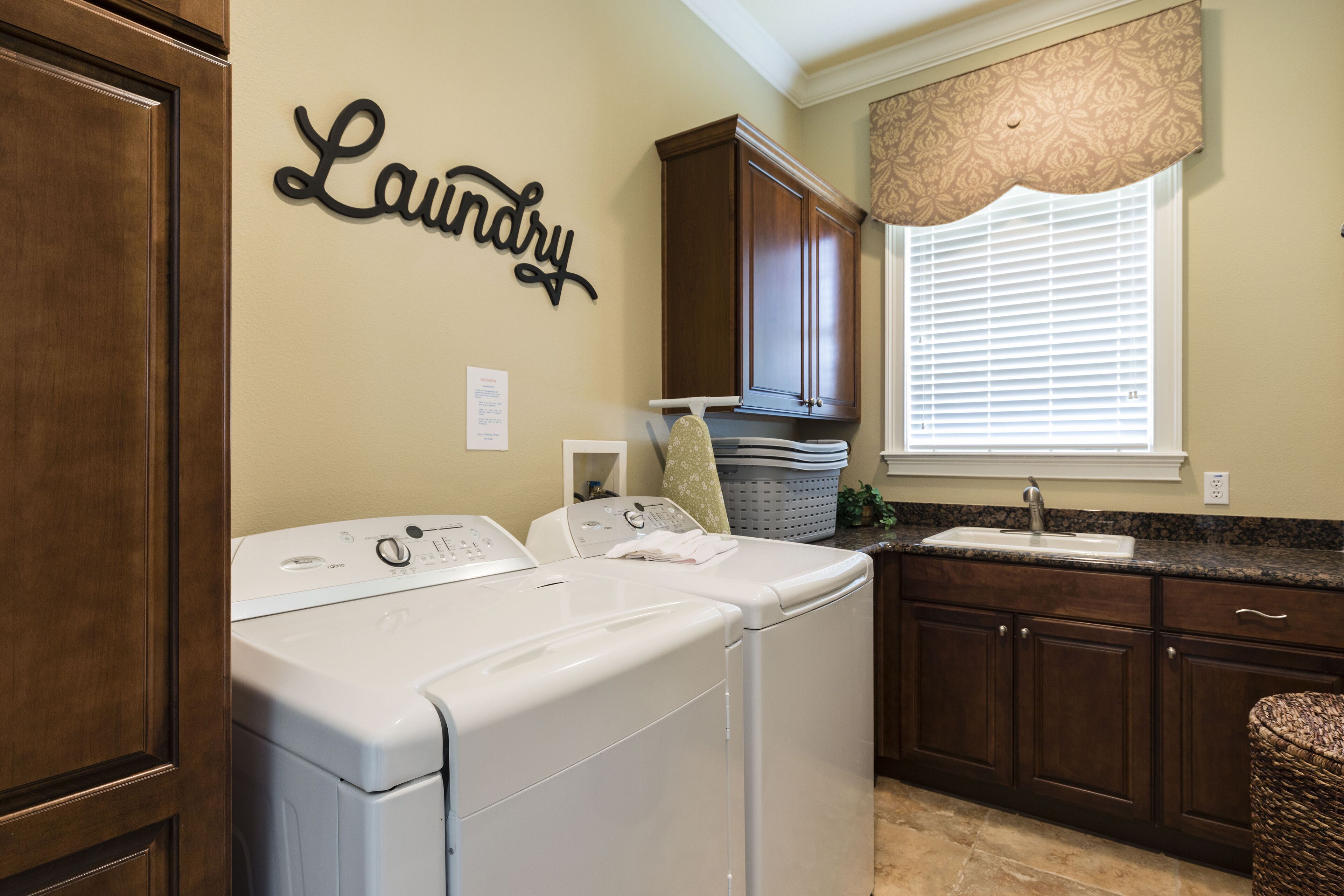 A laundry area with a wall-mounted sign, a washing machine, a dryer, and a window with blinds.