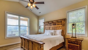 Une chambre à coucher avec un lit en bois, de grandes fenêtres équipées de stores, un ventilateur de plafond et une table de chevet avec une lampe.