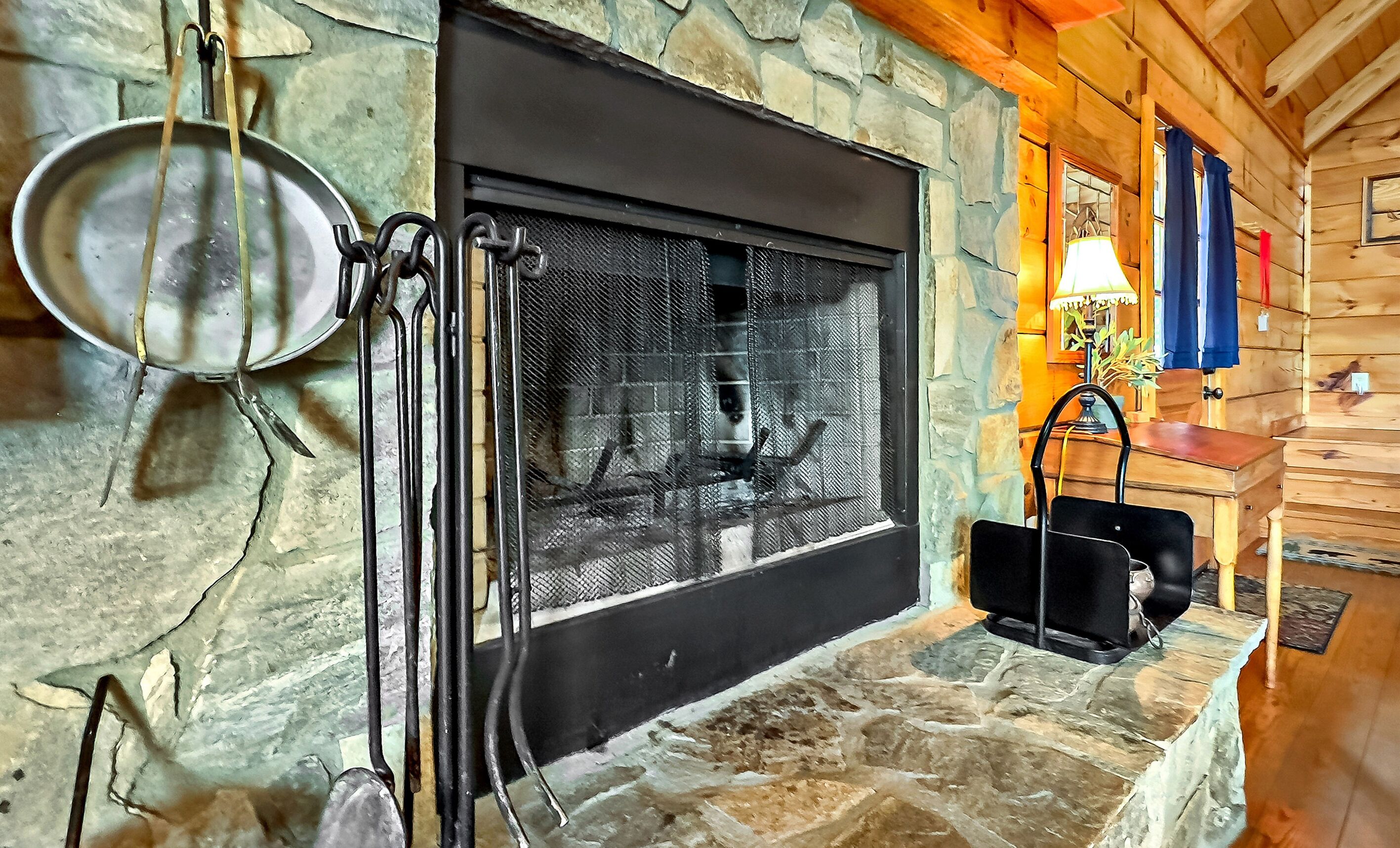 A stone fireplace with a metal screen, a hanging pot rack, and a wooden bench.