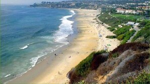 Una playa costera con una extensa franja de arena, algunos edificios al fondo y un paisaje montañoso.