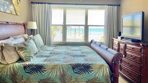 Une chambre à coucher avec un lit, un ventilateur de plafond, un téléviseur posé sur une commode et une fenêtre avec des rideaux.