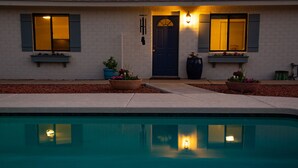 Une piscine avec une porte bleu foncé et des fenêtres dotées de volets, entourée d’un mur de briques blanches et de plantes en pot.
