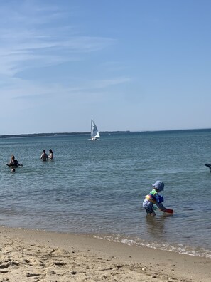 Una playa con gente en el agua y un velero a lo lejos.