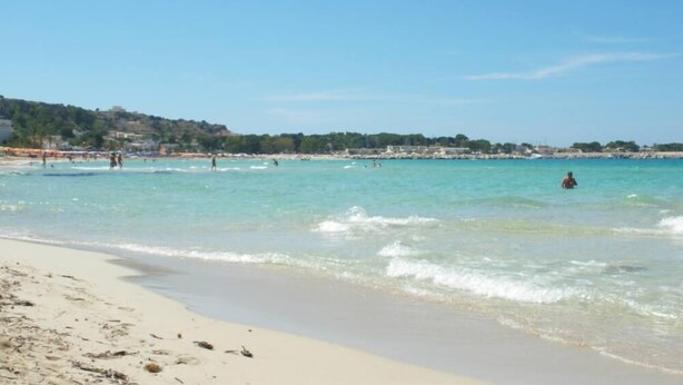 A beach with clear turquoise water, sandy shore, and a distant hill with buildings.