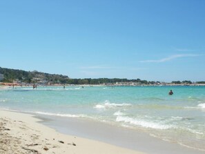 Ein Strand mit klarem türkisblauem Wasser, sandigem Ufer und einer entfernten Hügelkette mit Gebäuden.