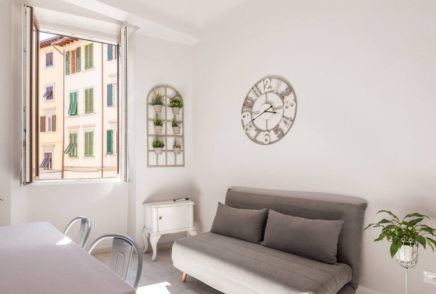 A bedroom with a bed, a bedside table, a wall clock, a window with a view of buildings, and a potted plant.