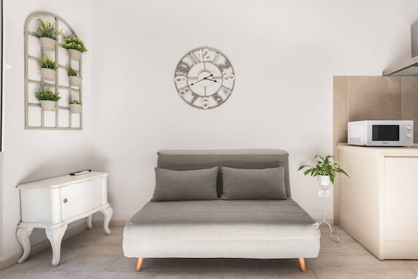 A bedroom with a grey sofa bed, a white bedside table, a white cabinet with a microwave, and a wall-mounted clock.