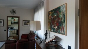 A living room with a red sofa, a table with a lamp, a painting, and a kitchen visible through an open door.