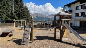 A playground with a slide and climbing structures, surrounded by a wooden fence and a building with a balcony.