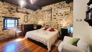 A bedroom with stone walls, a wooden floor, a bed with white bedding, a desk with a chair, and a window with curtains.