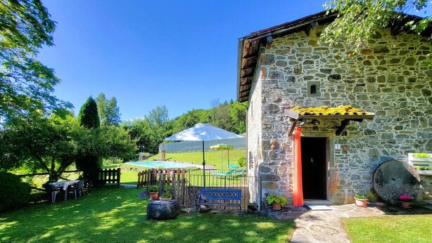 A stone building with a door, a bench, and a small garden area with a pool and umbrellas.