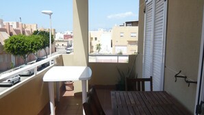 A balcony with a wooden table and chairs, a white table, and a view of buildings and a palm tree. - Nice apartment in Cabo de Gata (Cabo De Gata)