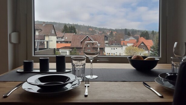 Une table à manger dressée avec des assiettes, des verres et des couverts, et par la fenêtre, on peut admirer des maisons et des arbres.