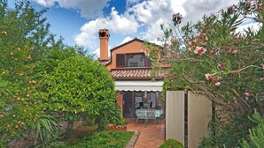 Ein Haus mit Ziegeldach, ein Garten mit grünen Sträuchern und rosa Blüten sowie eine Terrasse mit Tisch und Stühlen.