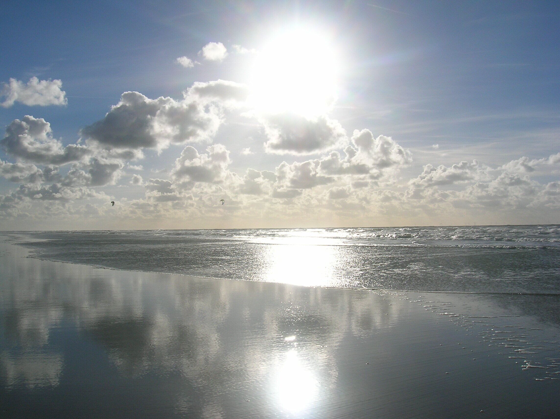 Une plage avec un ciel dégagé, une mer calme et le reflet du soleil sur le sable mouillé.