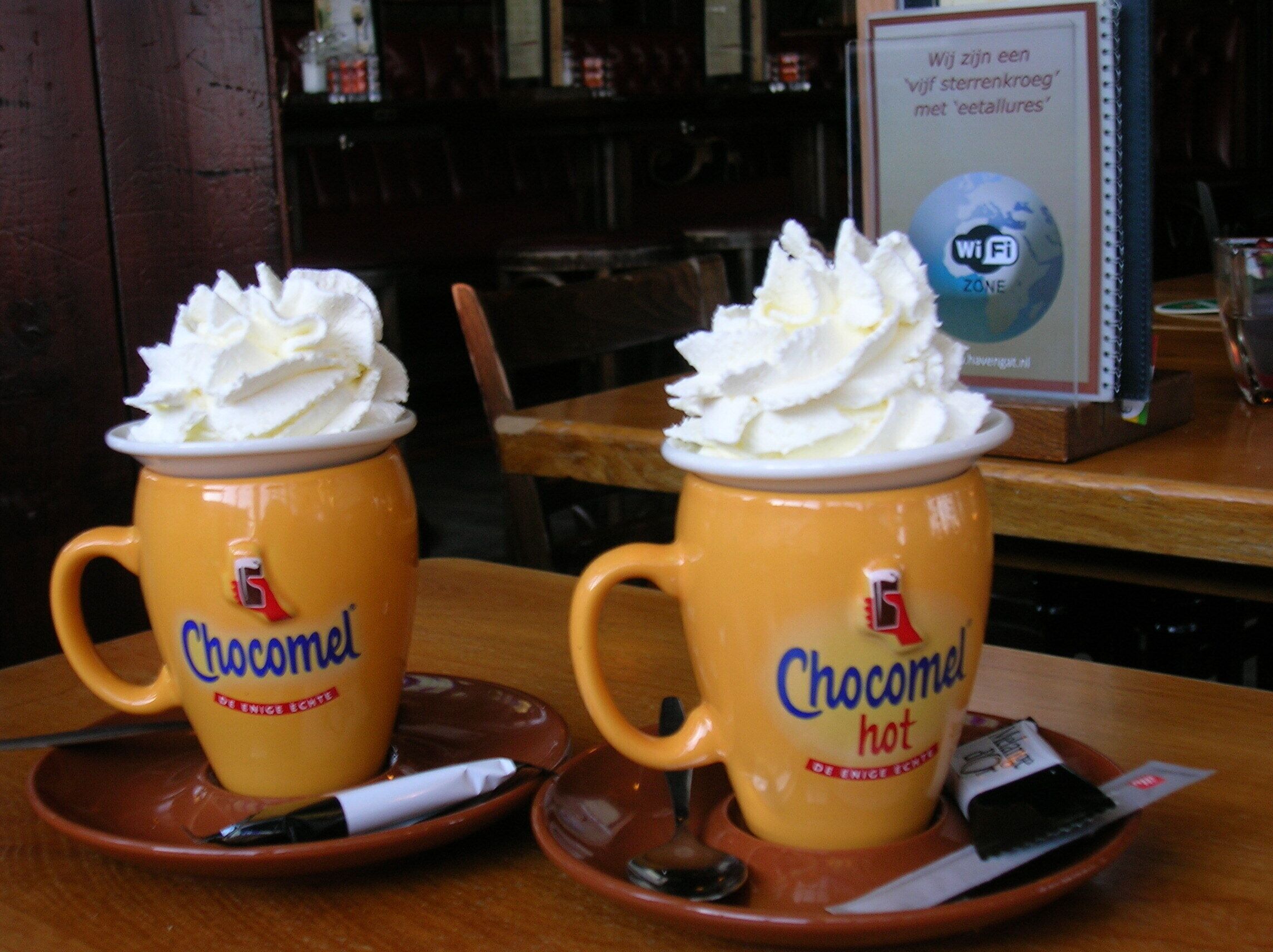 Two yellow mugs with whipped cream on a table, one labeled 'Chocomel' and the other 'Chocomel hot'.
