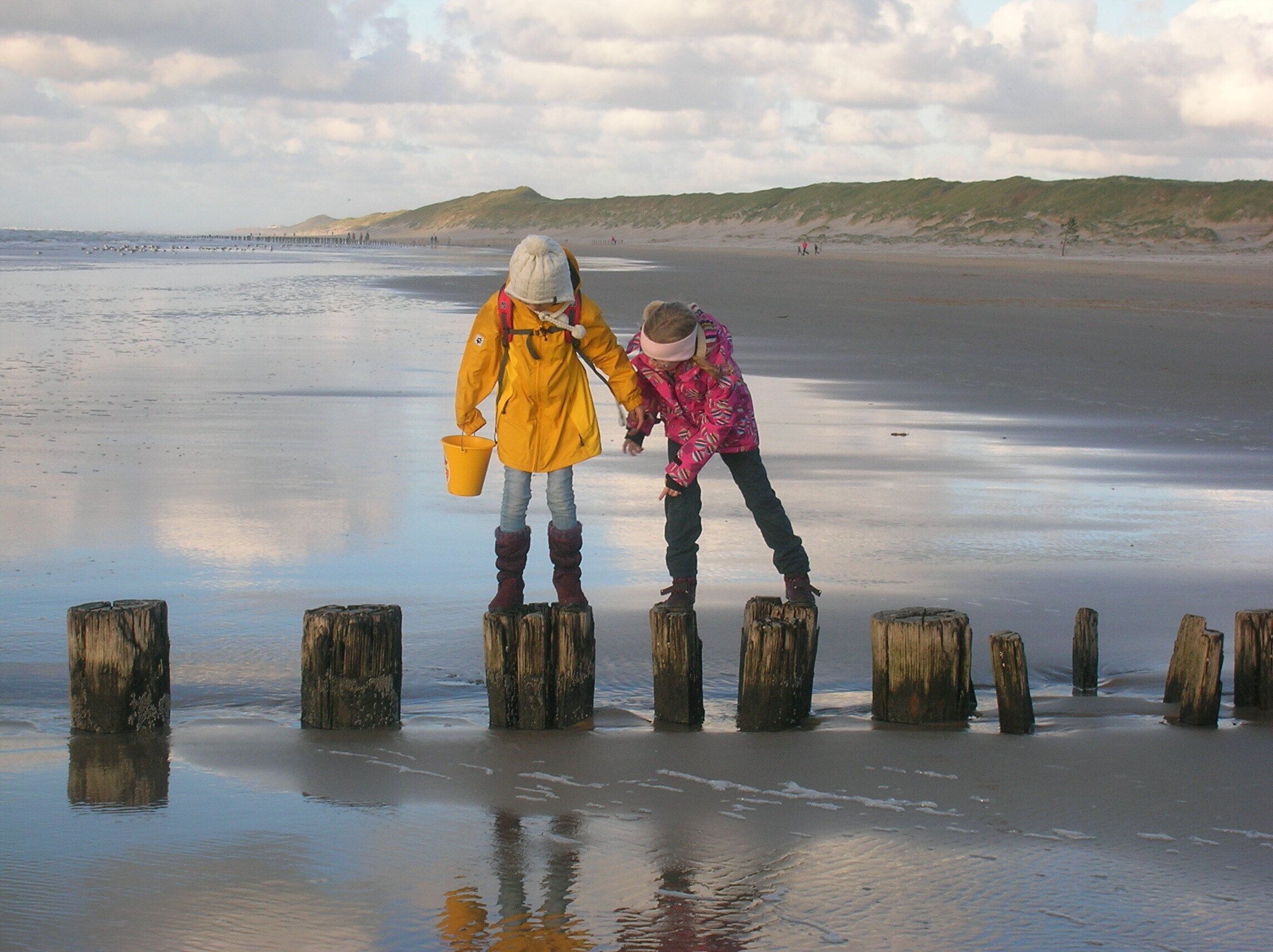 Two children playing on wooden posts in a sandy area with a beach and dunes in the background.