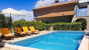 A swimming pool with lounge chairs and an umbrella, surrounded by greenery and a building with a tiled roof.