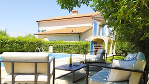 A poolside area with lounge chairs and a table, a clear view of a house with a tiled roof and blue shutters, and a well-maintained hedge in the background.