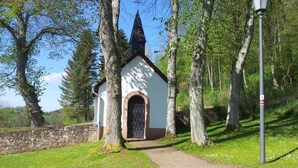 A small white church with a dark roof and arched entrance, surrounded by trees and a stone wall. - big holiday house with grassland in the Vulkaneifel, near Nürburgring and Maare (Lissendorf)