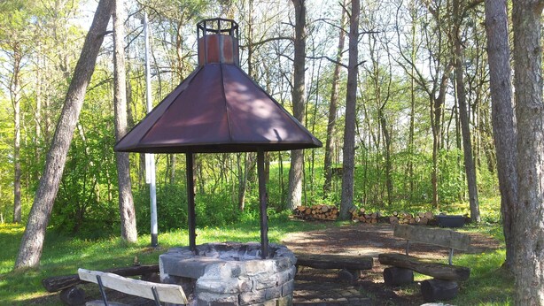 A rustic outdoor seating area with a stone fire pit, wooden benches, and a metal gazebo. - big holiday house with grassland in the Vulkaneifel, near Nürburgring and Maare (Lissendorf)