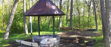 A rustic outdoor seating area with a stone fire pit, wooden benches, and a metal gazebo.