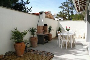 Een terras buiten met een barbecue, planten in potten en een witte tafel met stoelen.
