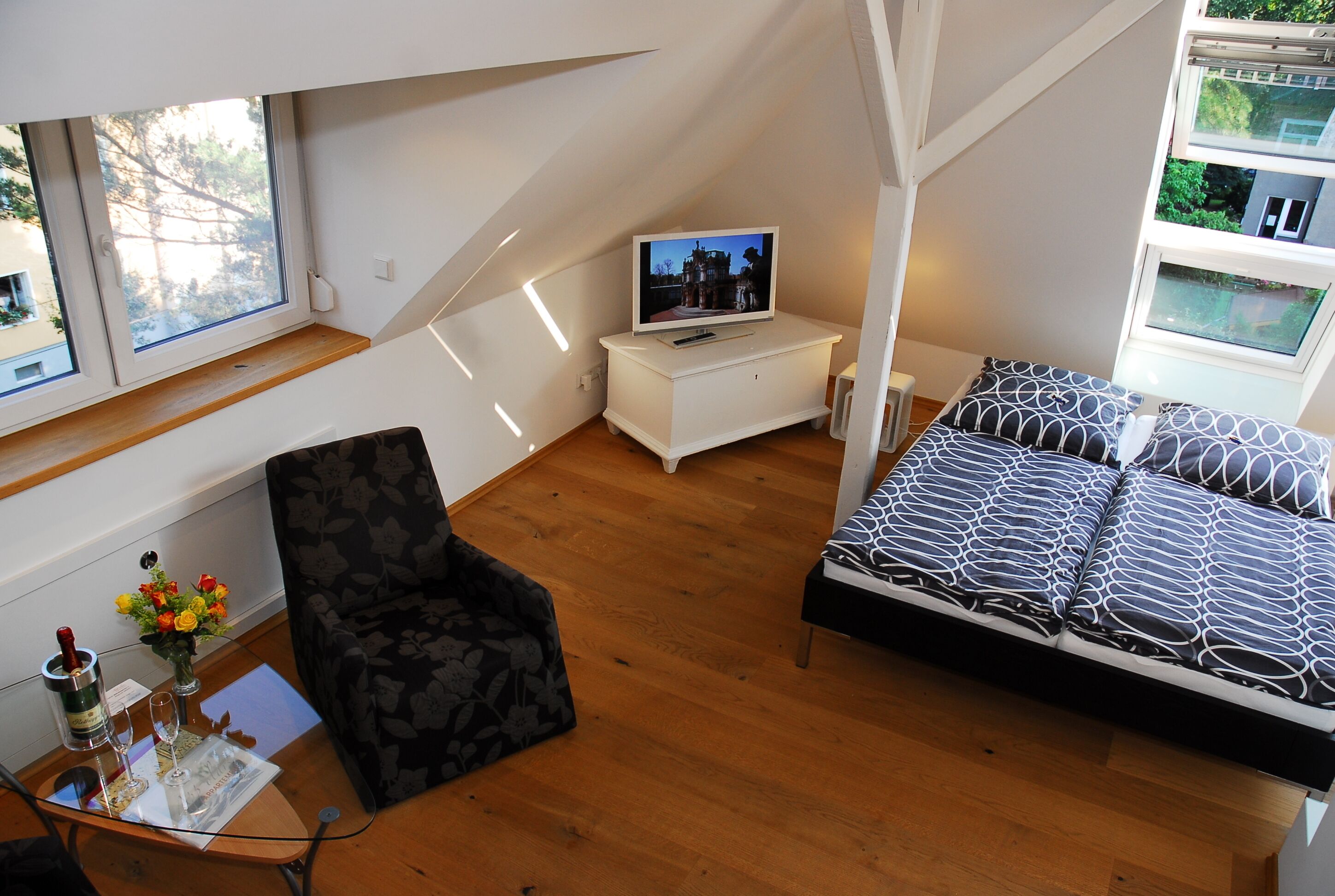 A small, cozy room with a bed, a chair, a TV, and a small table with a bottle and glasses.