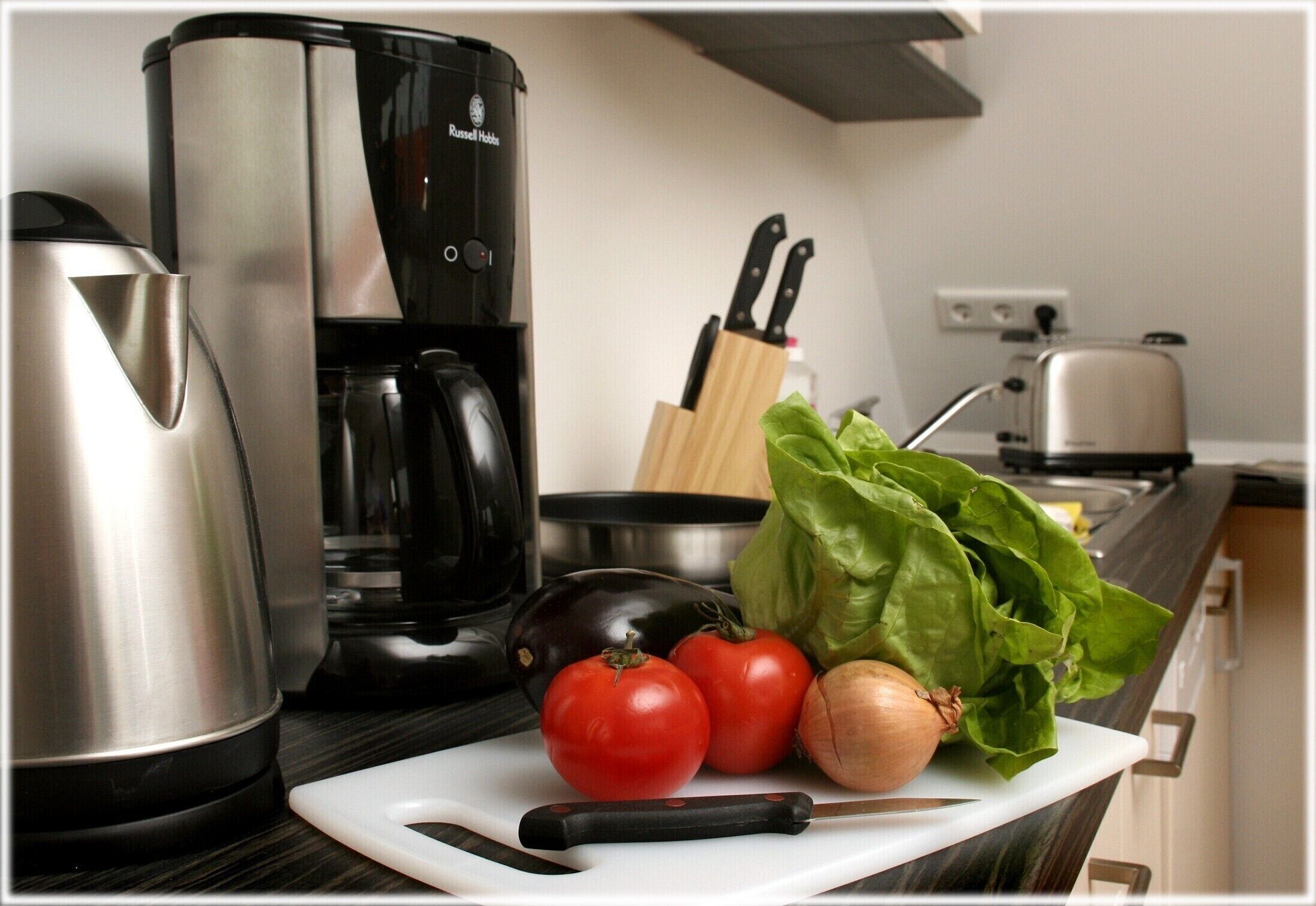 Un plan de travail de cuisine avec une machine à café, une bouilloire, une planche à découper avec des légumes et un grille-pain.